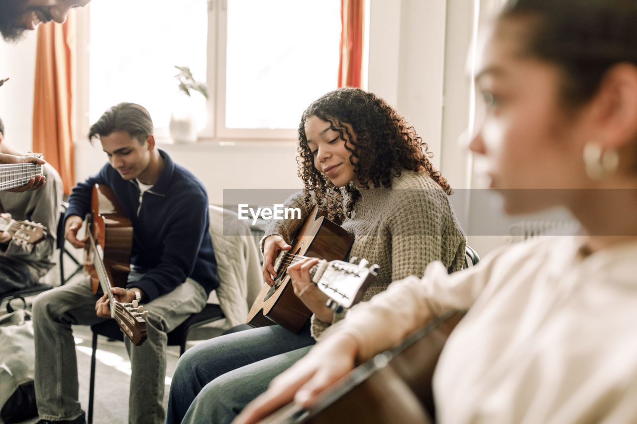 Smiling teenage girl practicing guitar with friends in music class at high school