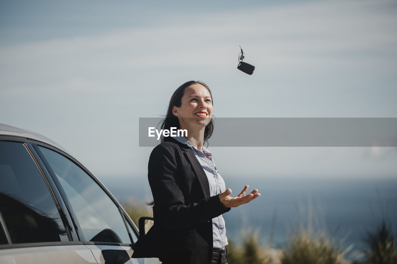 Smiling entrepreneur playing with car key while standing against sky