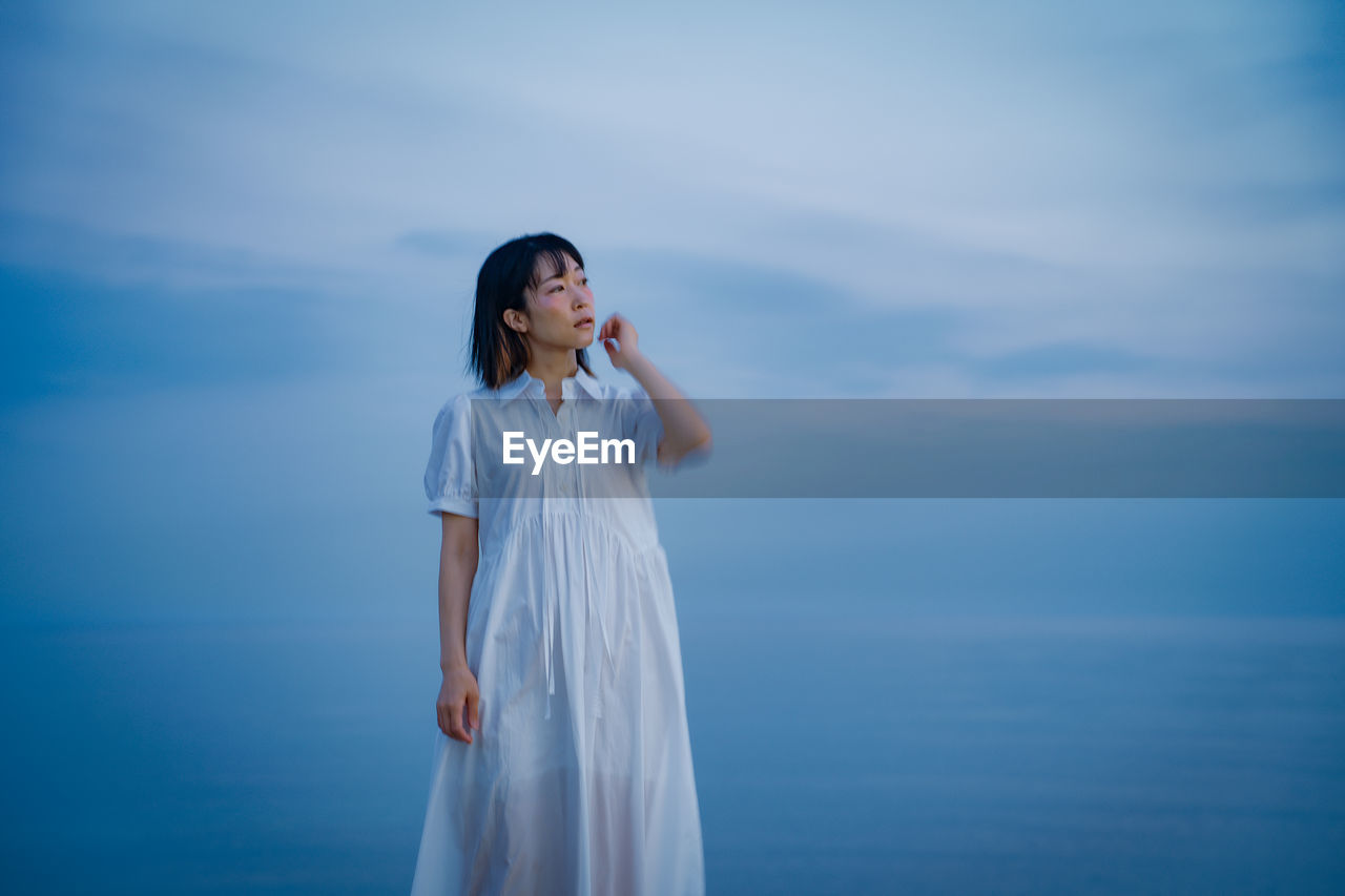 blue, one person, sky, women, young adult, standing, adult, water, nature, sea, long hair, dress, hairstyle, white, cloud, fashion, horizon, clothing, sunlight, looking, contemplation, person, tranquility, copy space, outdoors, beauty in nature, three quarter length, emotion, bride, female, reflection, religion, spirituality