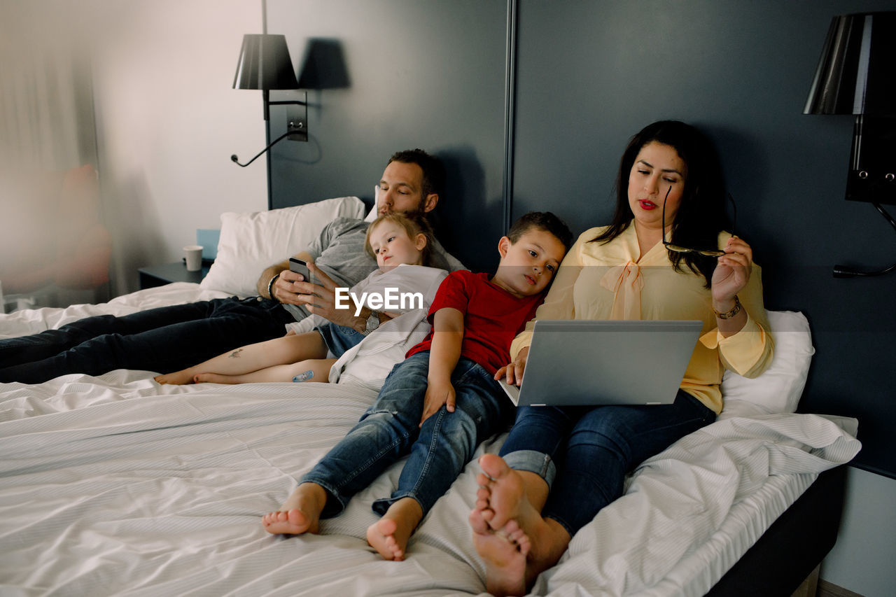 Parents using technologies while sitting with children on bed in bedroom