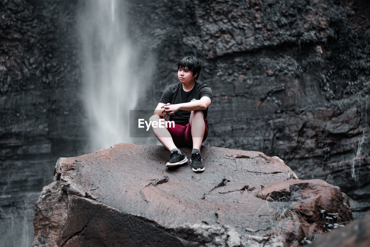 Full length of man sitting on rock against waterfall