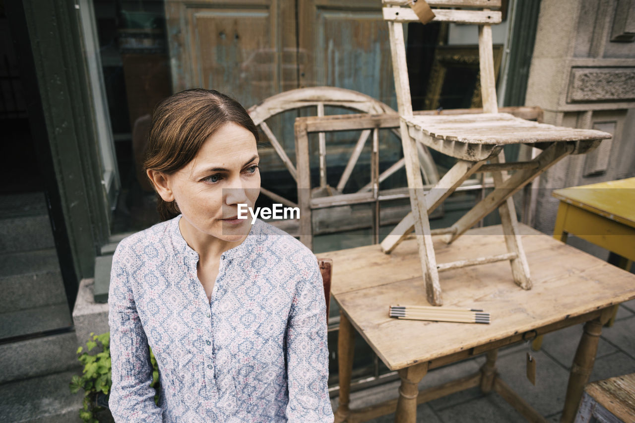 Thoughtful woman sitting by tables and chairs against store