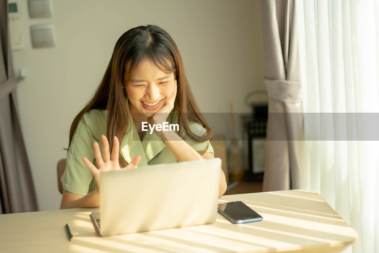 PORTRAIT OF SMILING YOUNG WOMAN USING SMART PHONE IN LAPTOP