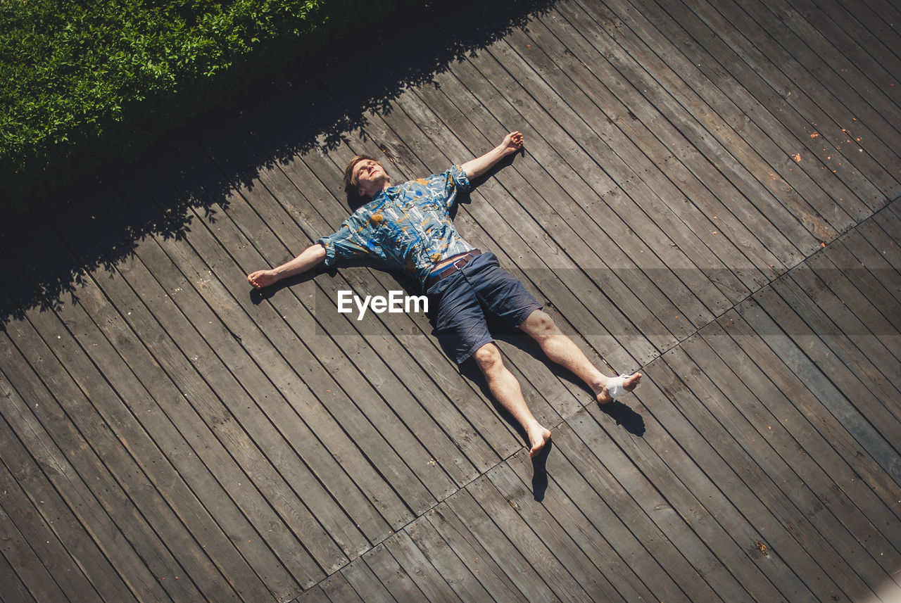 High angle view of carefree man with arms outstretched lying on boardwalk