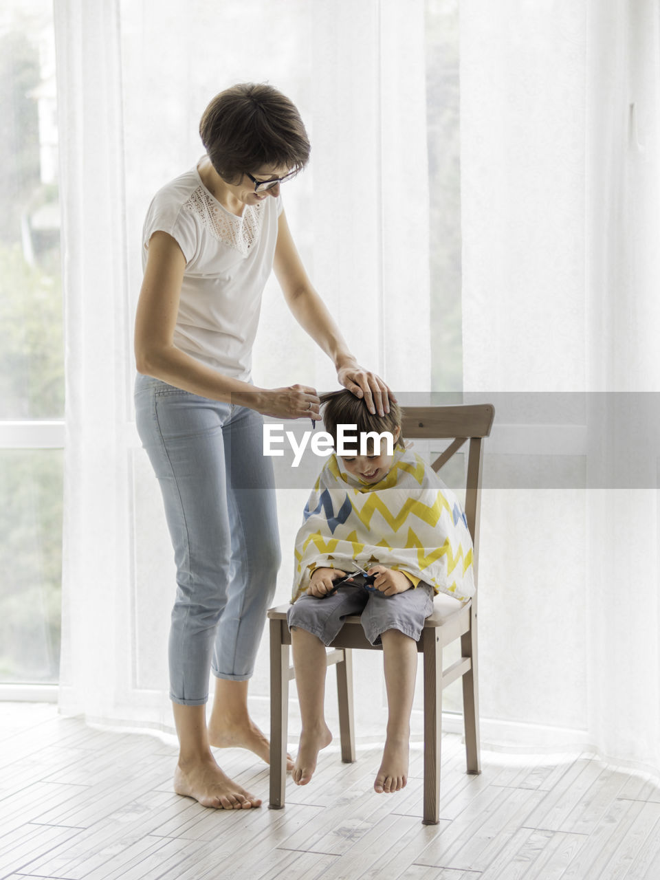 Mother cuts her son's hair by herself. little boy sits, covered with cloth. new normal.