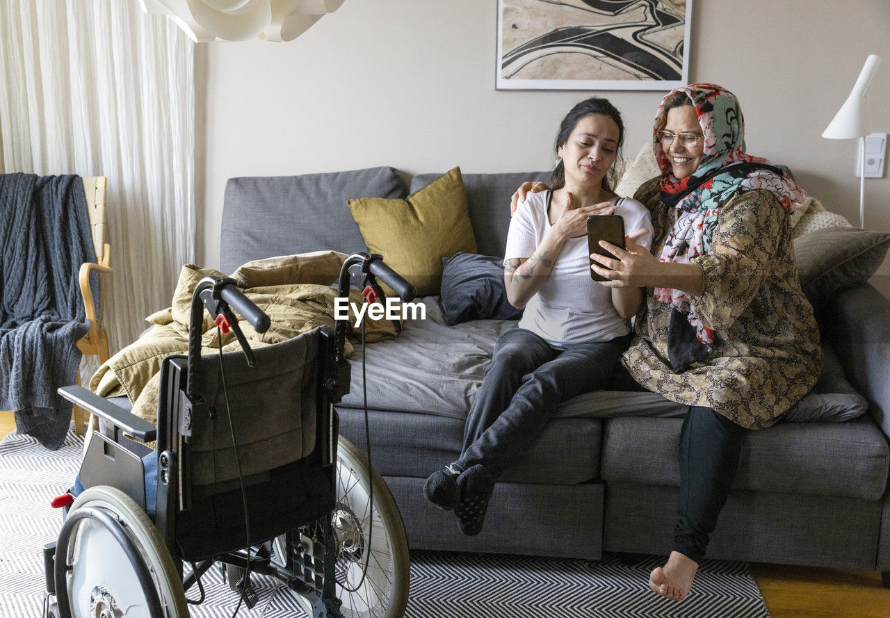 Woman with paraplegia doing video call through smart phone while sitting with mother on sofa at home