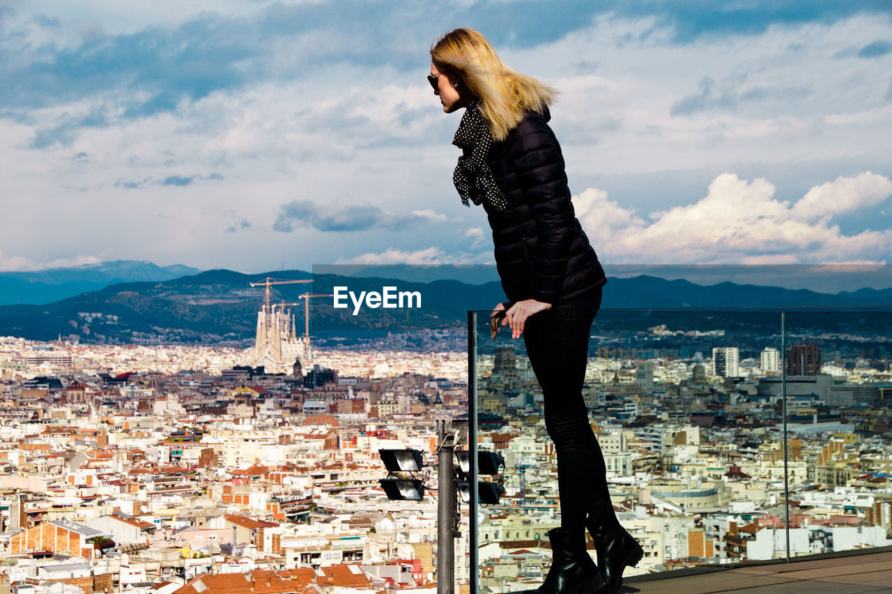 Young woman wearing warm clothing while standing on balcony against cityscape