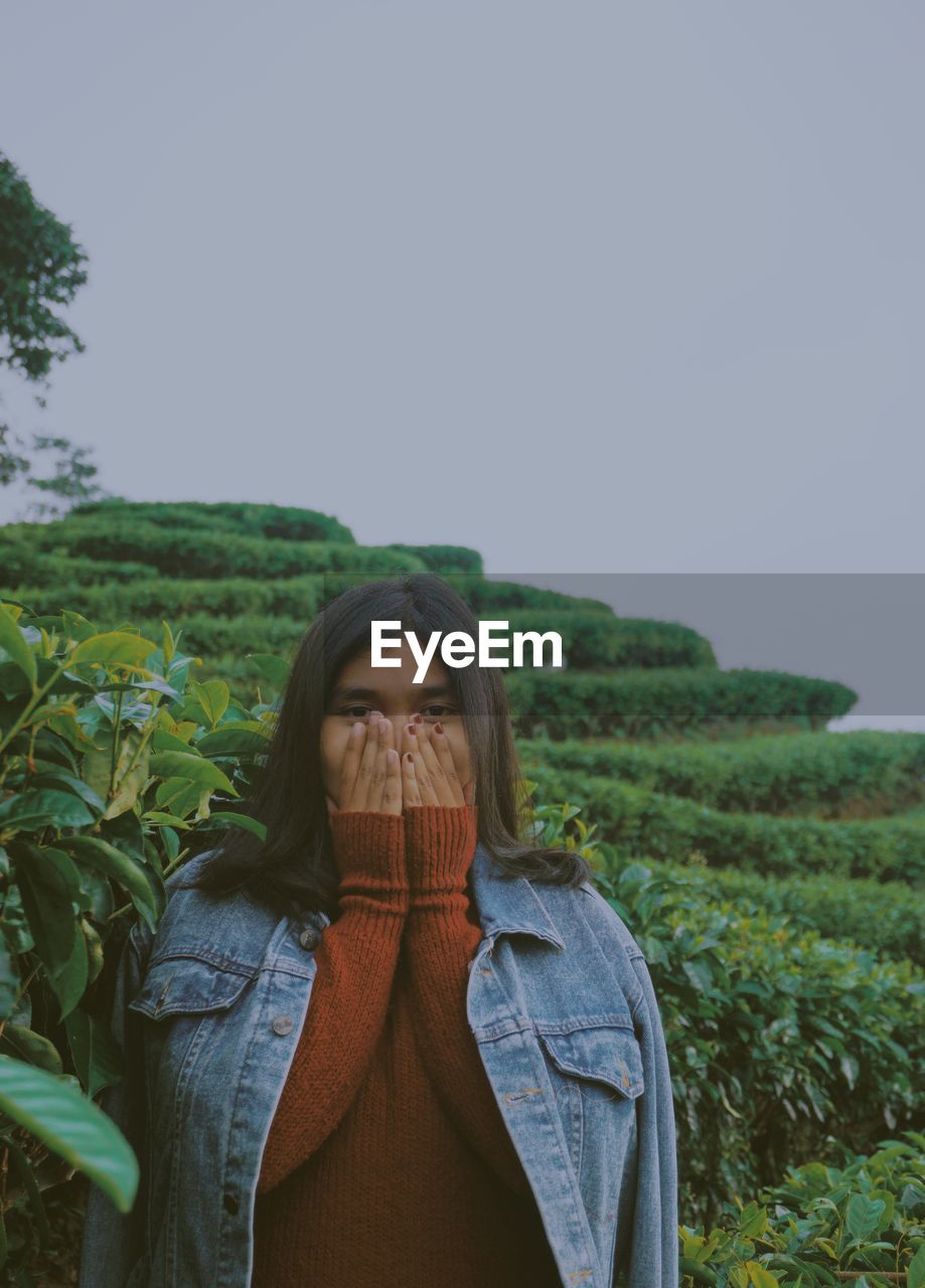 Woman covering mouth while standing against plants and sky