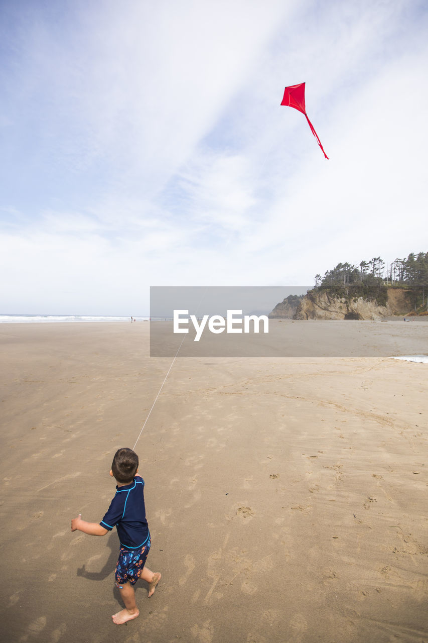 Young boy looks up at red kite flying in the sky at the beach.