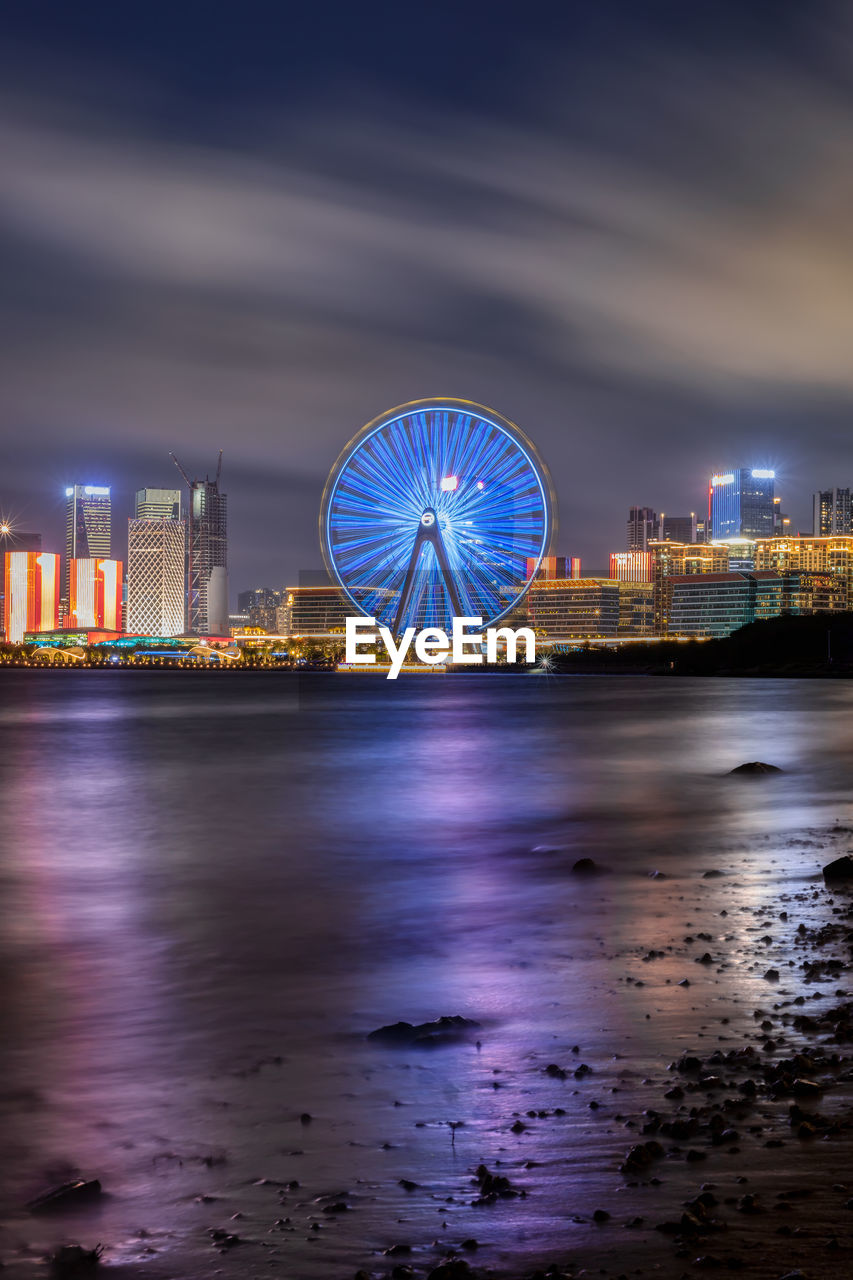 ILLUMINATED FERRIS WHEEL BY SEA AGAINST SKY