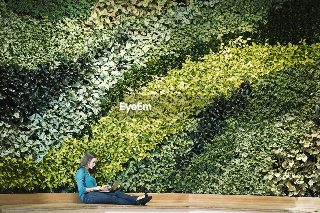 Young woman using laptop in front of green plant wall