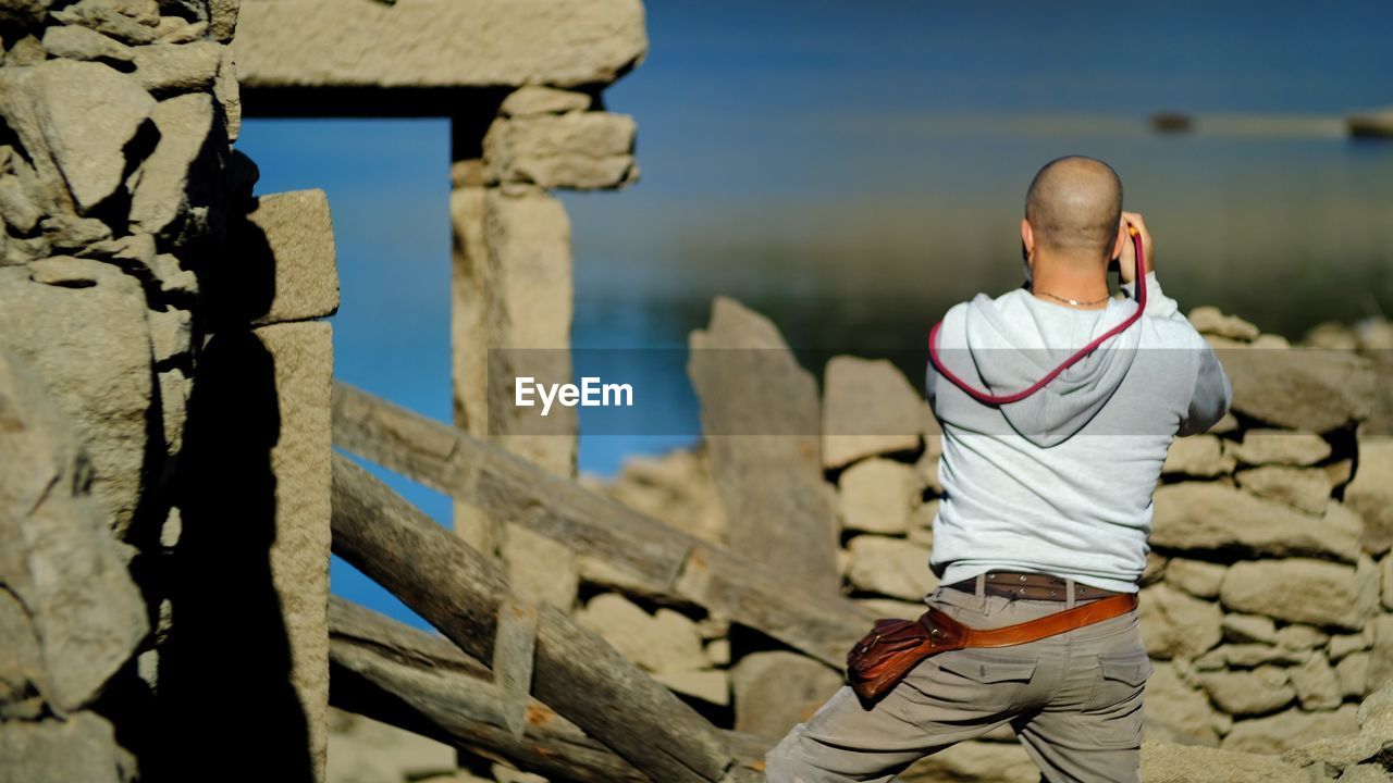 Rear view of man standing by stone wall during sunny day