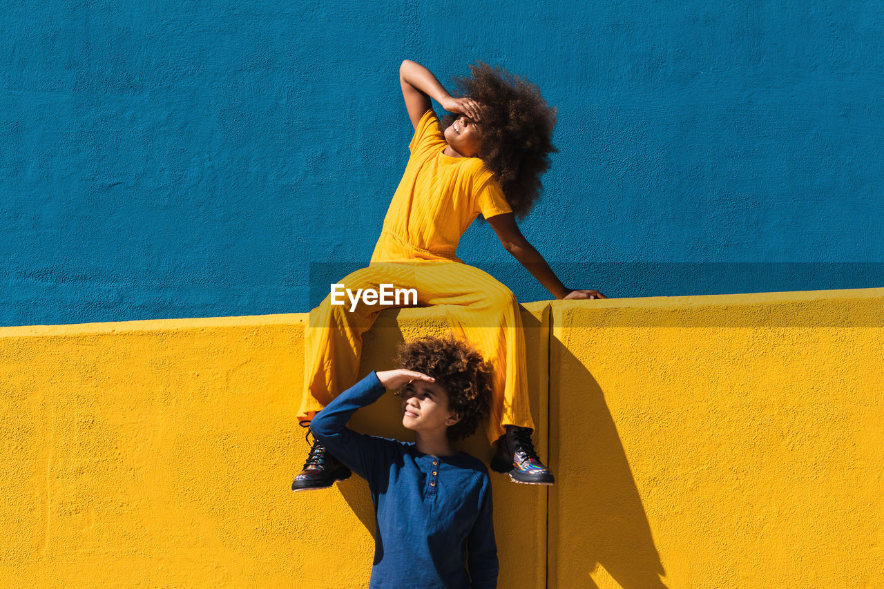 Teen african american girl and boy with afro hairstyle wearing colorful clothes resting together against blue and yellow wall in sunny day on street