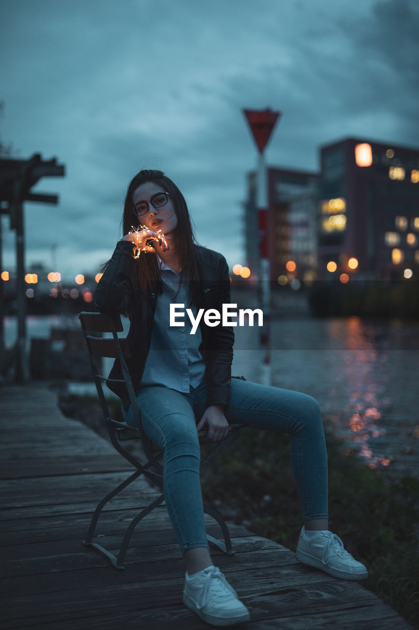 Portrait of young woman holding string light while sitting on bench by river against sky at dusk