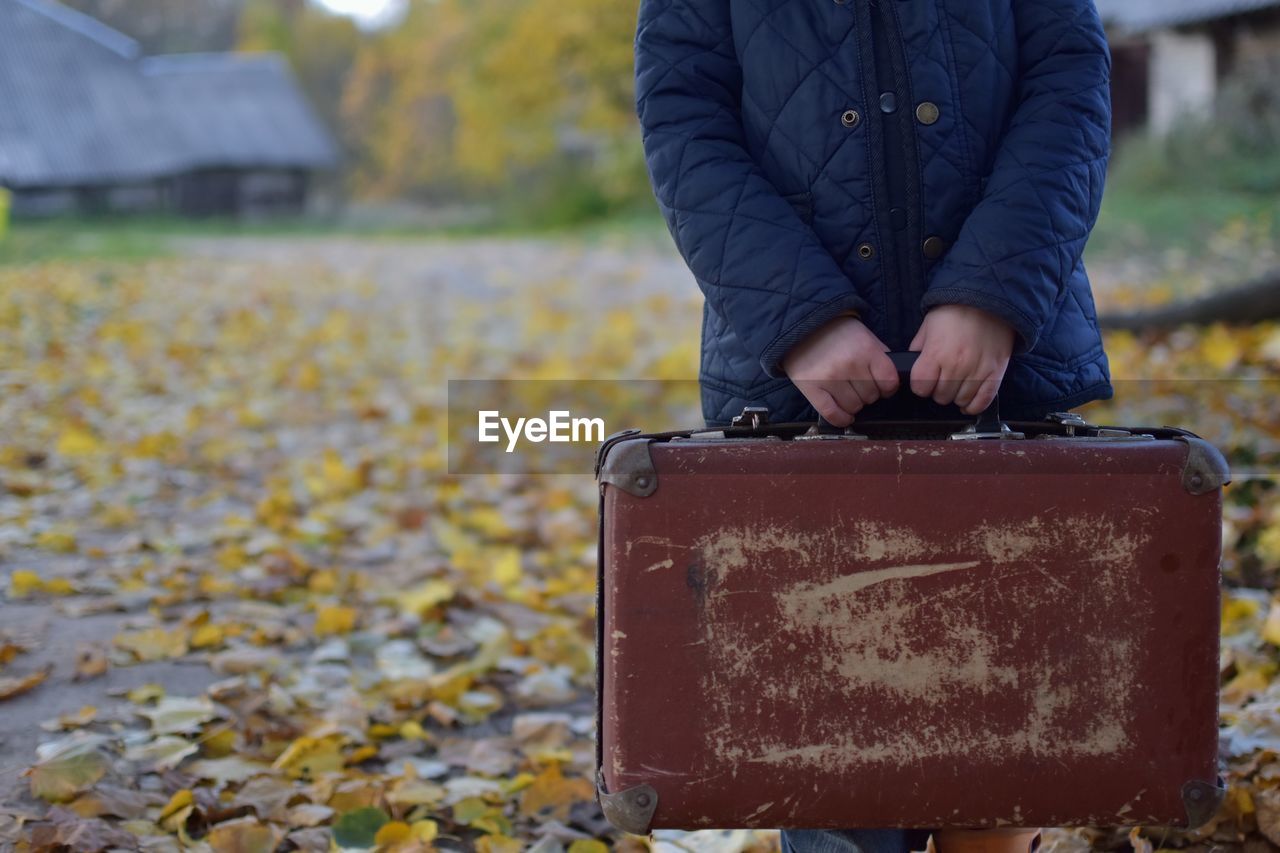Midsection of child holding old luggage while standing on field during autumn