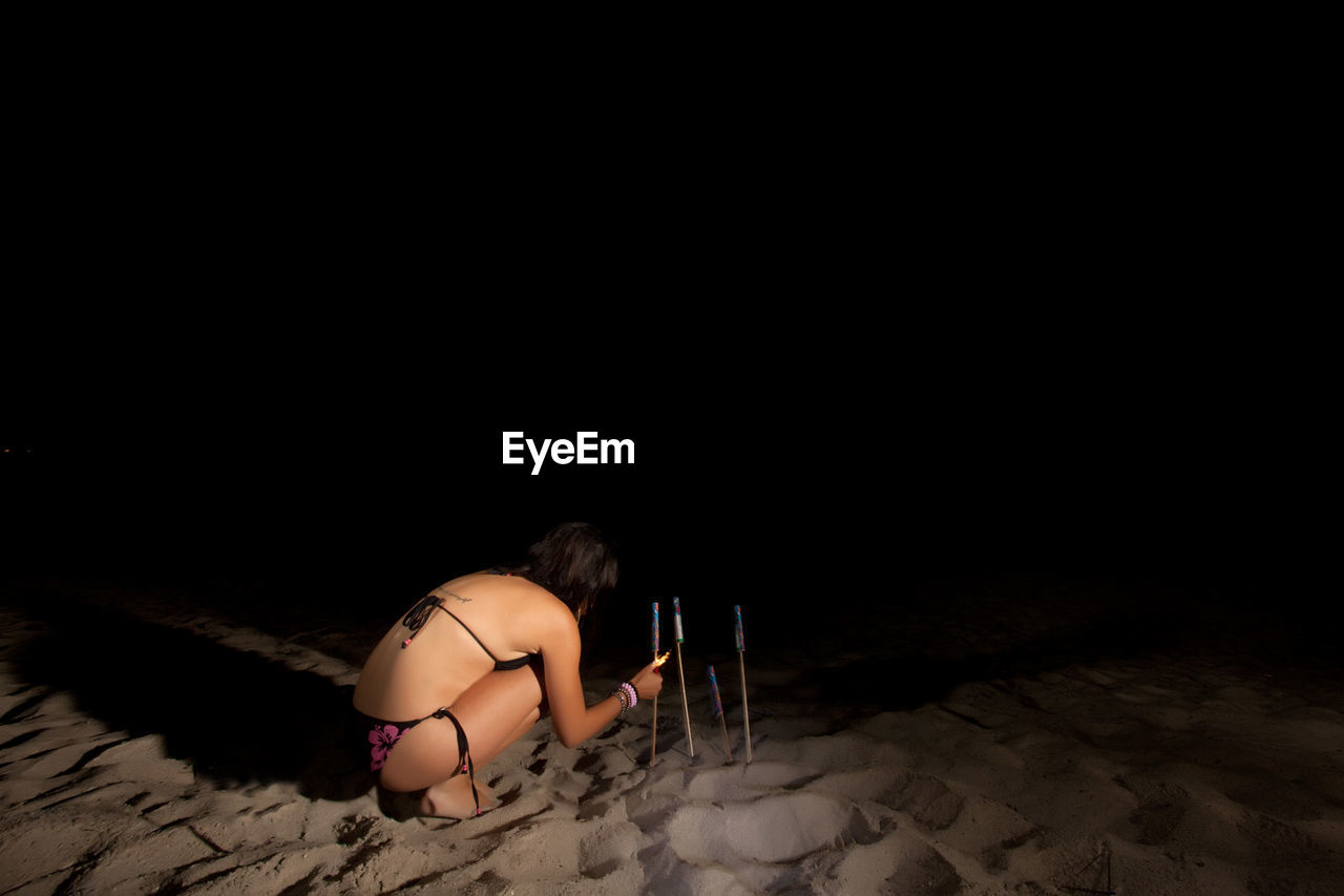 Rear view of woman wearing bikini while igniting rocket at beach during night