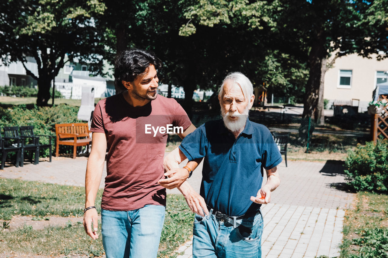 Smiling male caretaker looking while walking arm in arm with senior man at nursing home