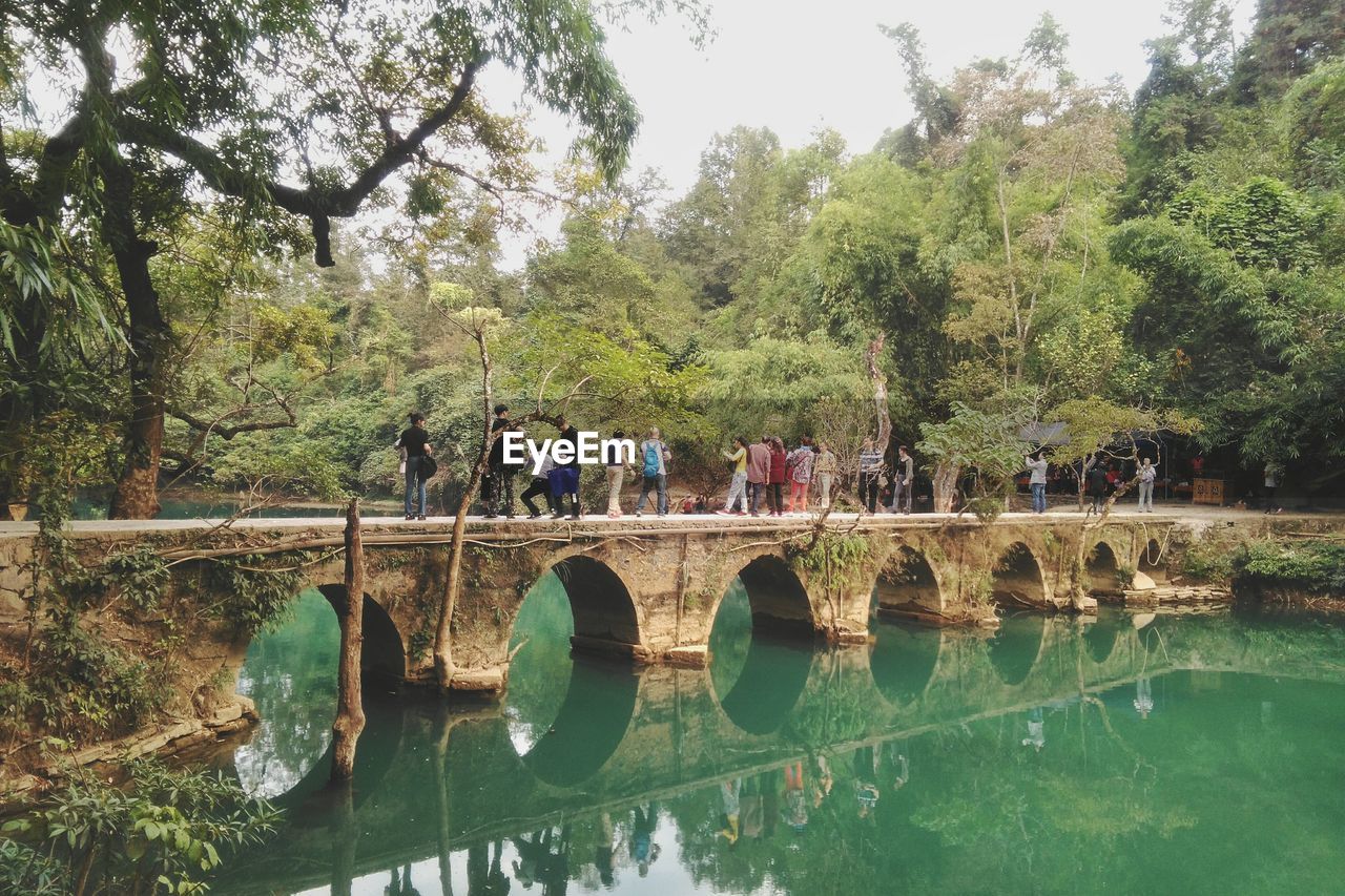 People on arch bridge with reflection in river against trees