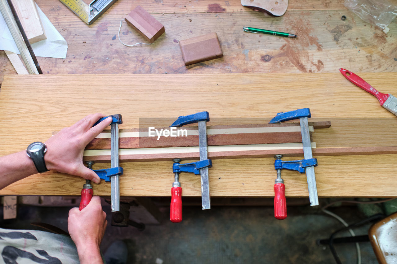 Overhead view of crop unrecognizable male carpenter fixing wooden sticks glued together with clamps at workbench with professional tools