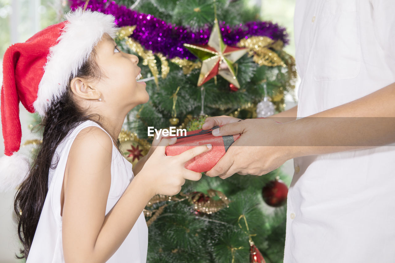 Midsection of man giving gift to girl against christmas tree at home