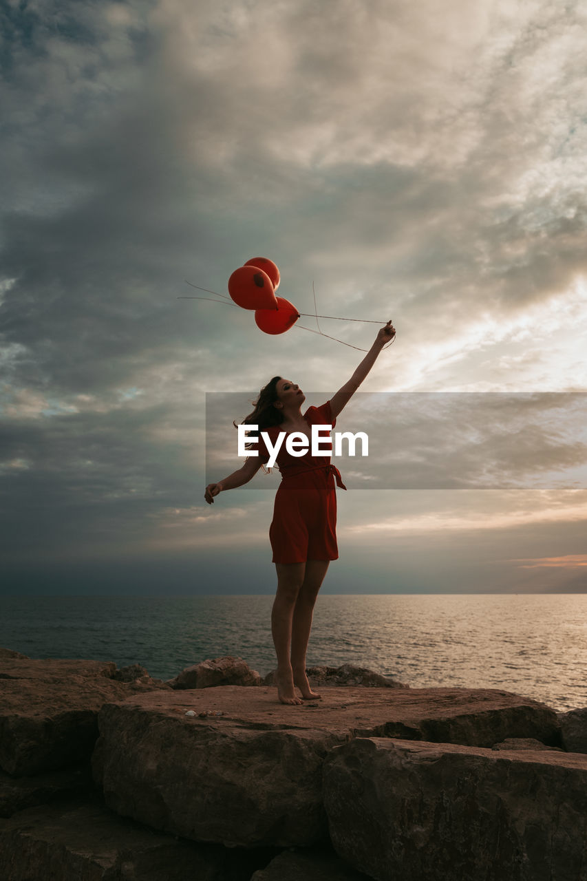 Woman holding balloons while standing on rock by sea against sky during sunset