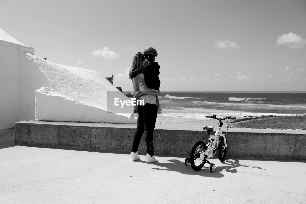 Rear view of mother carrying daughter while standing by bicycle against sea