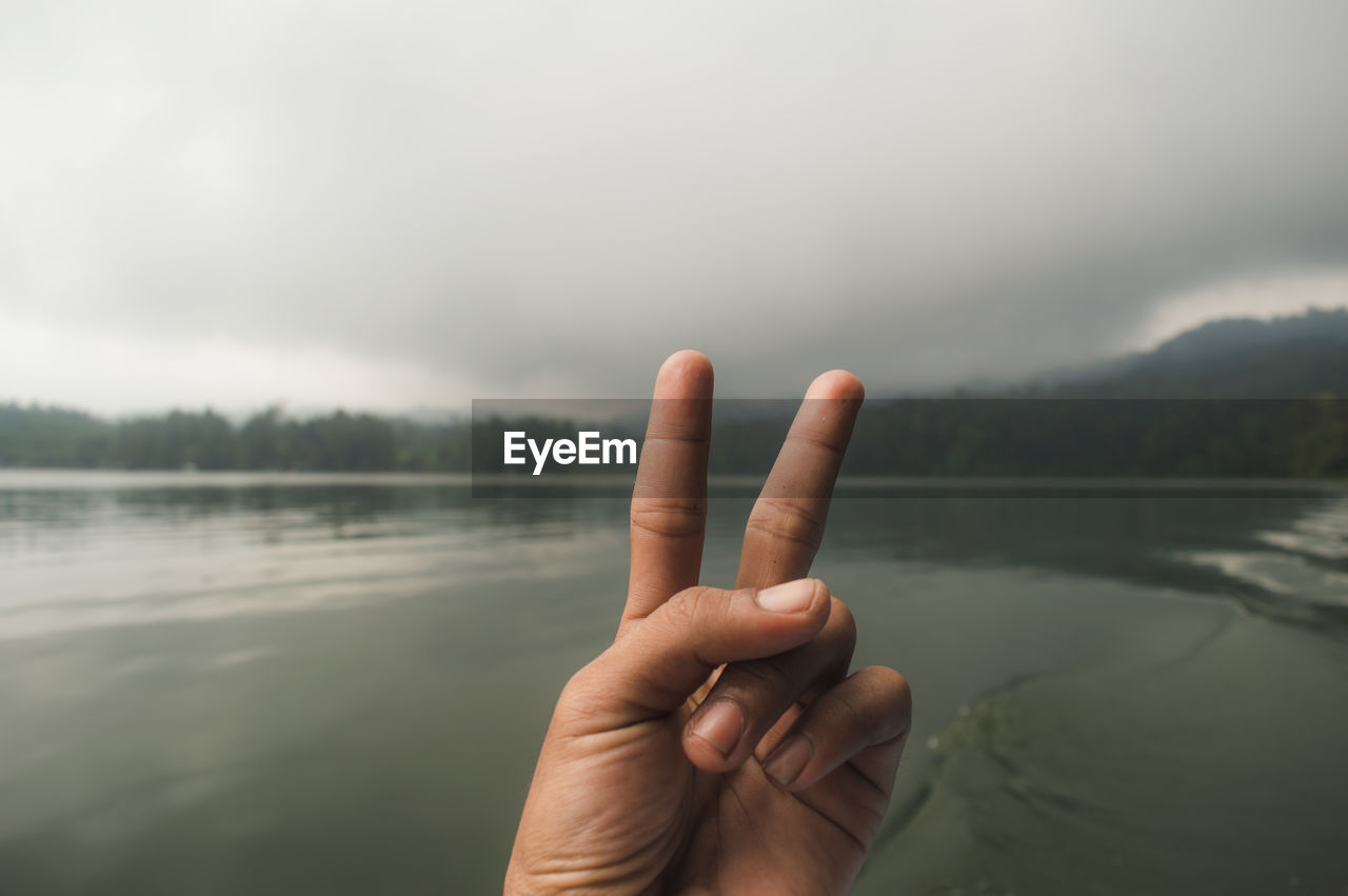 Cropped hand gesturing peace sign by lake against cloudy sky