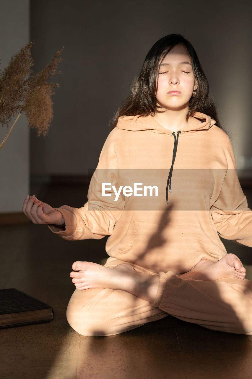 A girl in a beige tracksuit sits on the floor in a lotus position and meditates