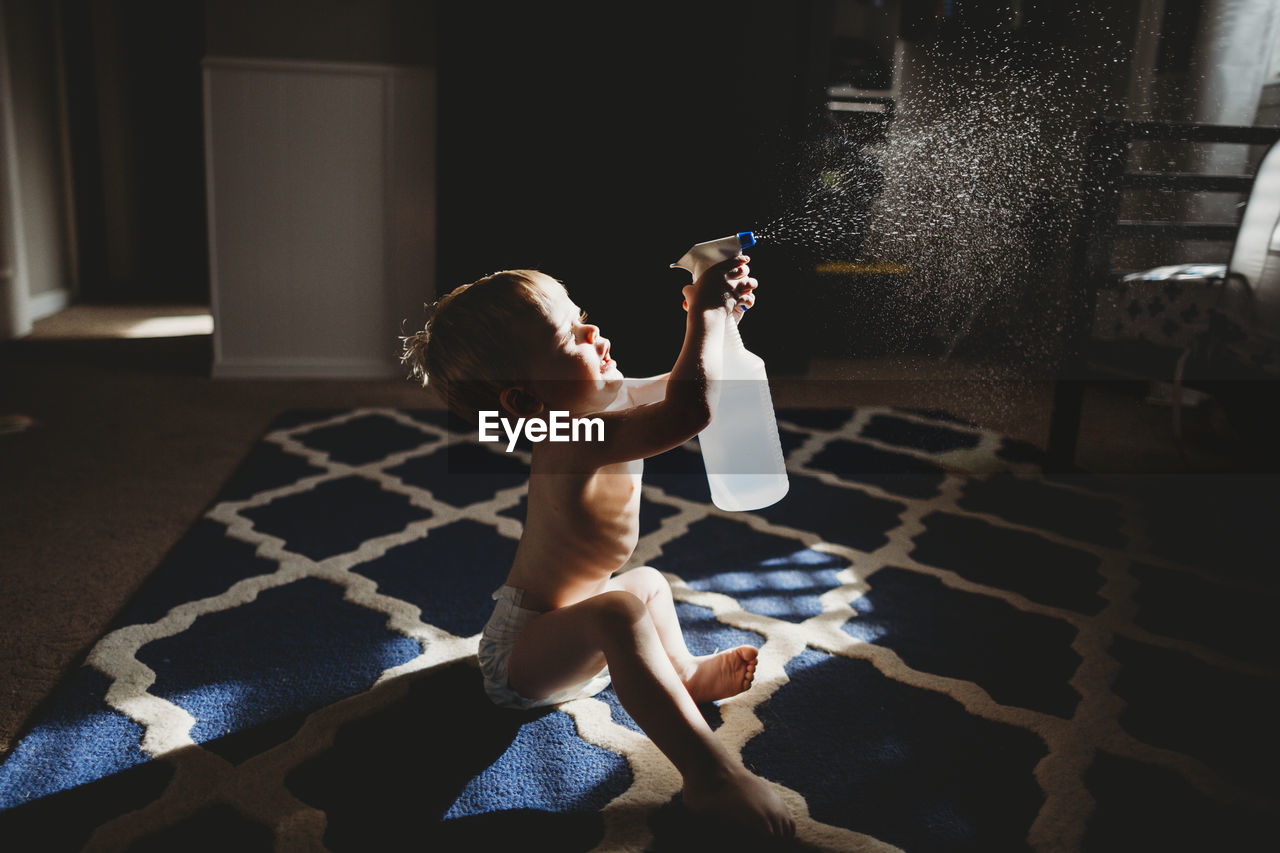 Side view of shirtless baby boy spraying water while sitting on carpet at home