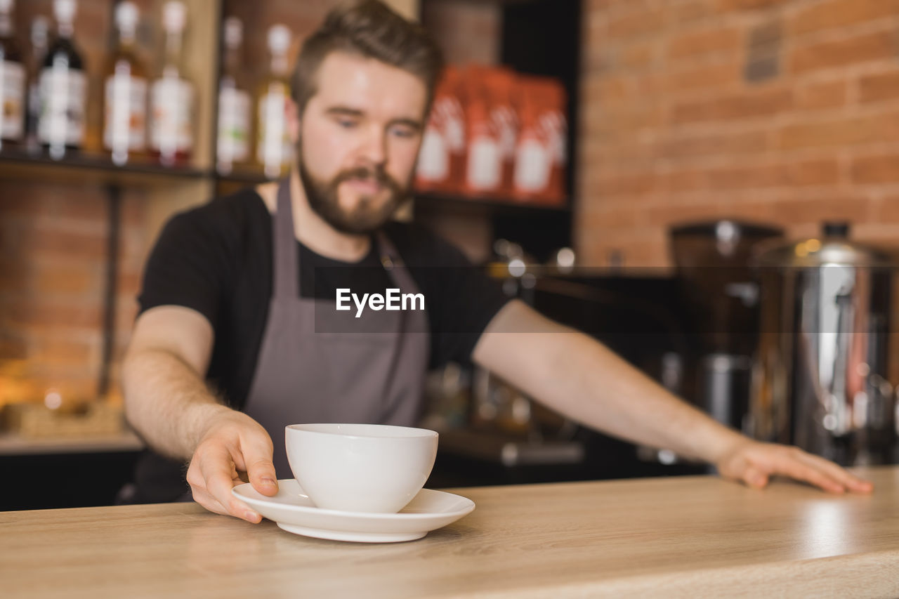 Bearded male barista in apron serving cup of coffee while working at bar table in coffee shop near coffee machine