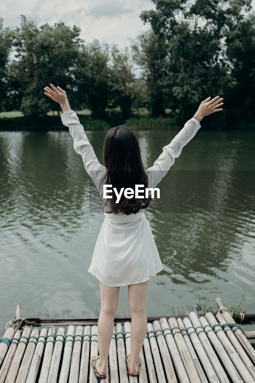 Rear view of woman with arms raised standing at lakeshore against tree