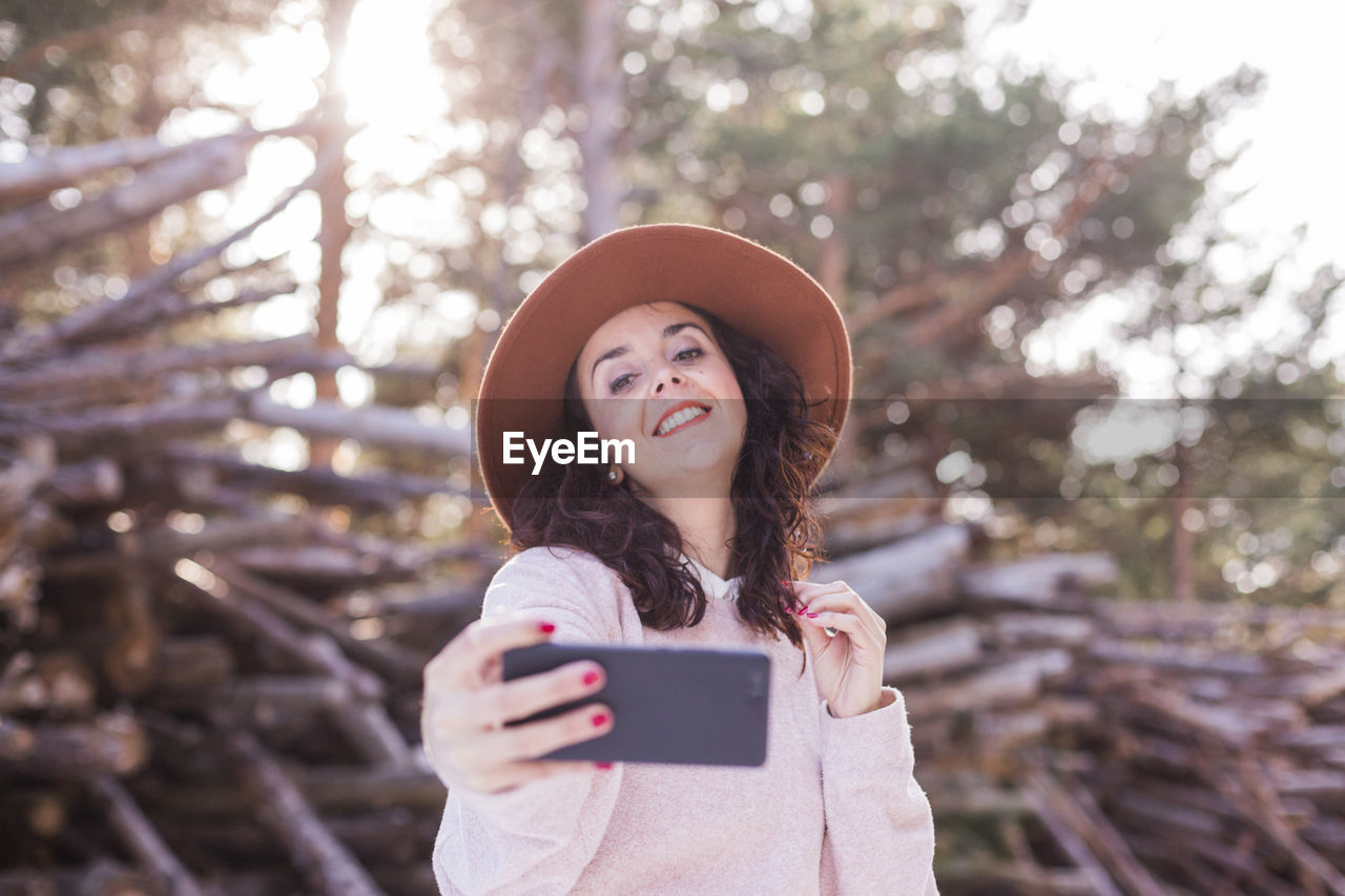 Smiling woman using smart phone while standing against woodpile in forest