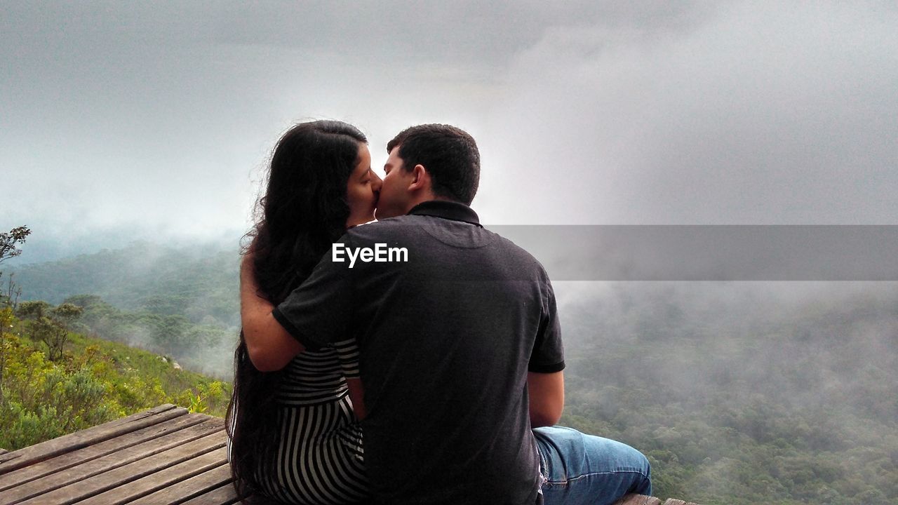 Rear view of young couple kissing while sitting on platform against fog