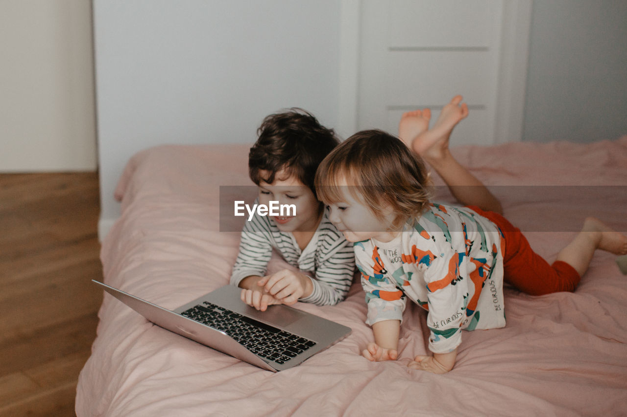 Little brother and sister lie in the bedroom on the bed and look at the laptop. high quality photo