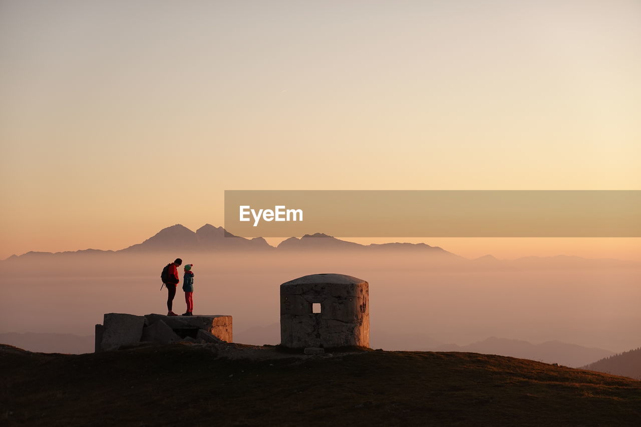 Father and daughter standing on rock against mountain and sky during sunset