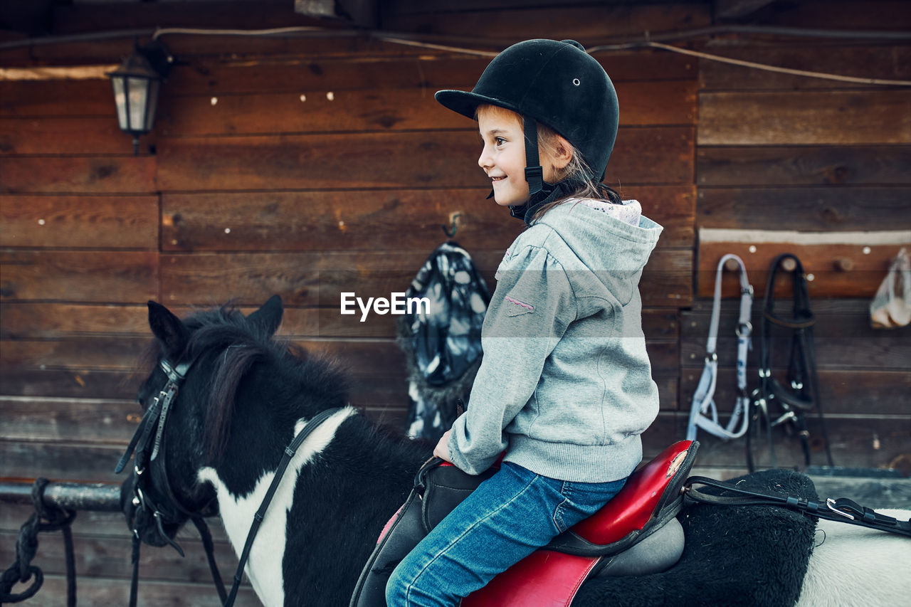 Little smiling girl learning horseback riding. 5-6 years old equestrian in helmet having fun riding