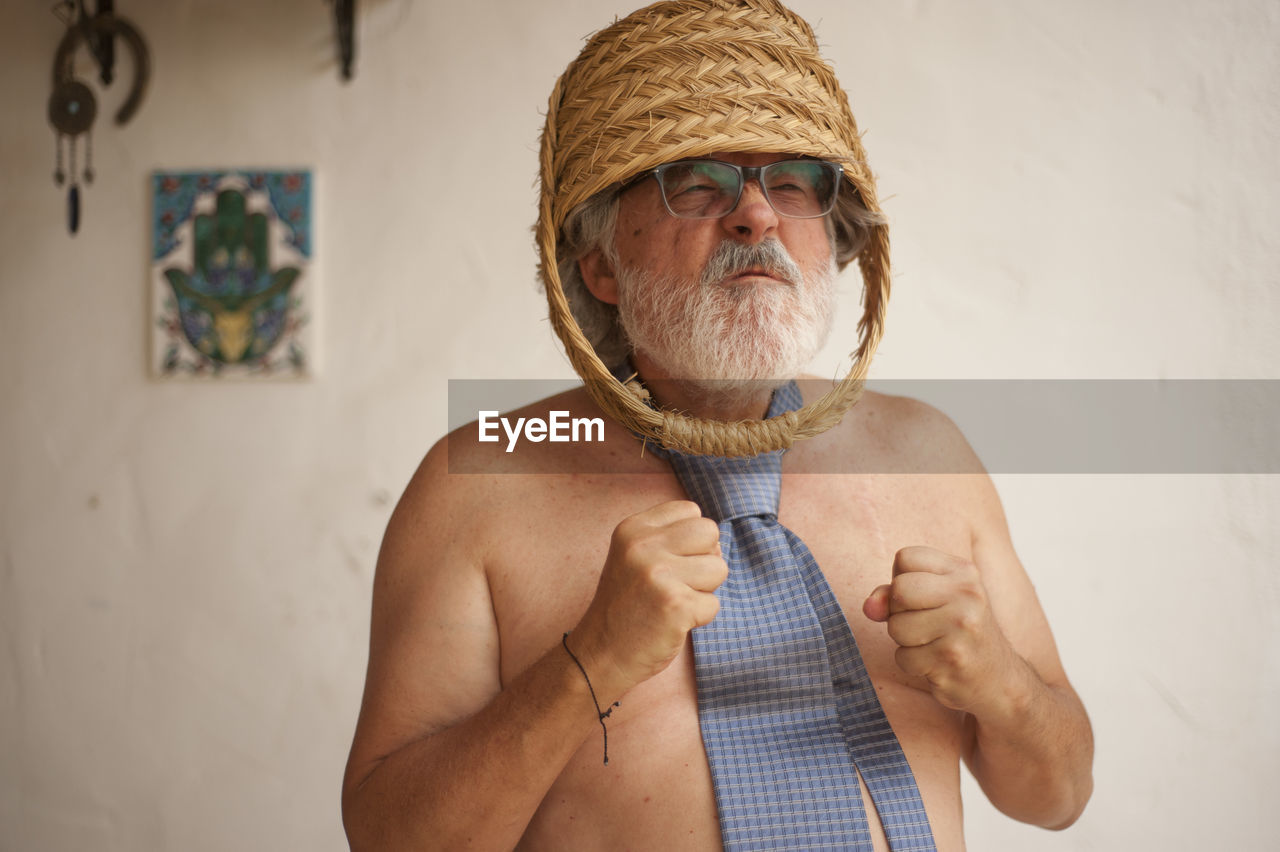 Shirtless man wearing basket while standing against wall