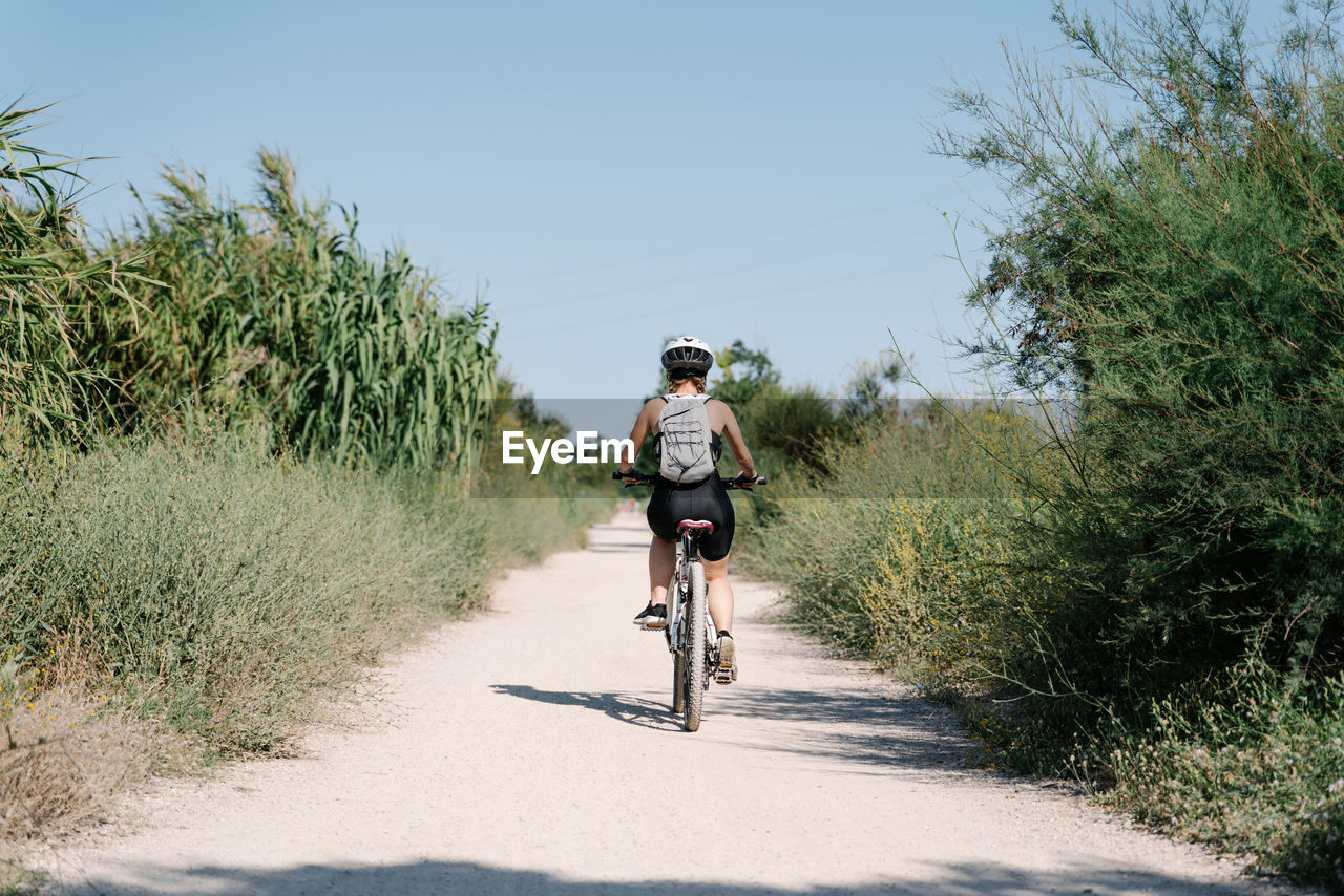 Full body of positive young female cyclist in sportswear and helmet riding bicycle on dirt path among green plants in summer day in countryside