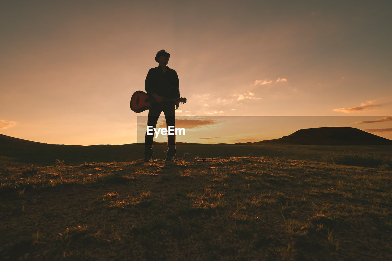 Silhouette man with guitar standing on field against sky during sunset