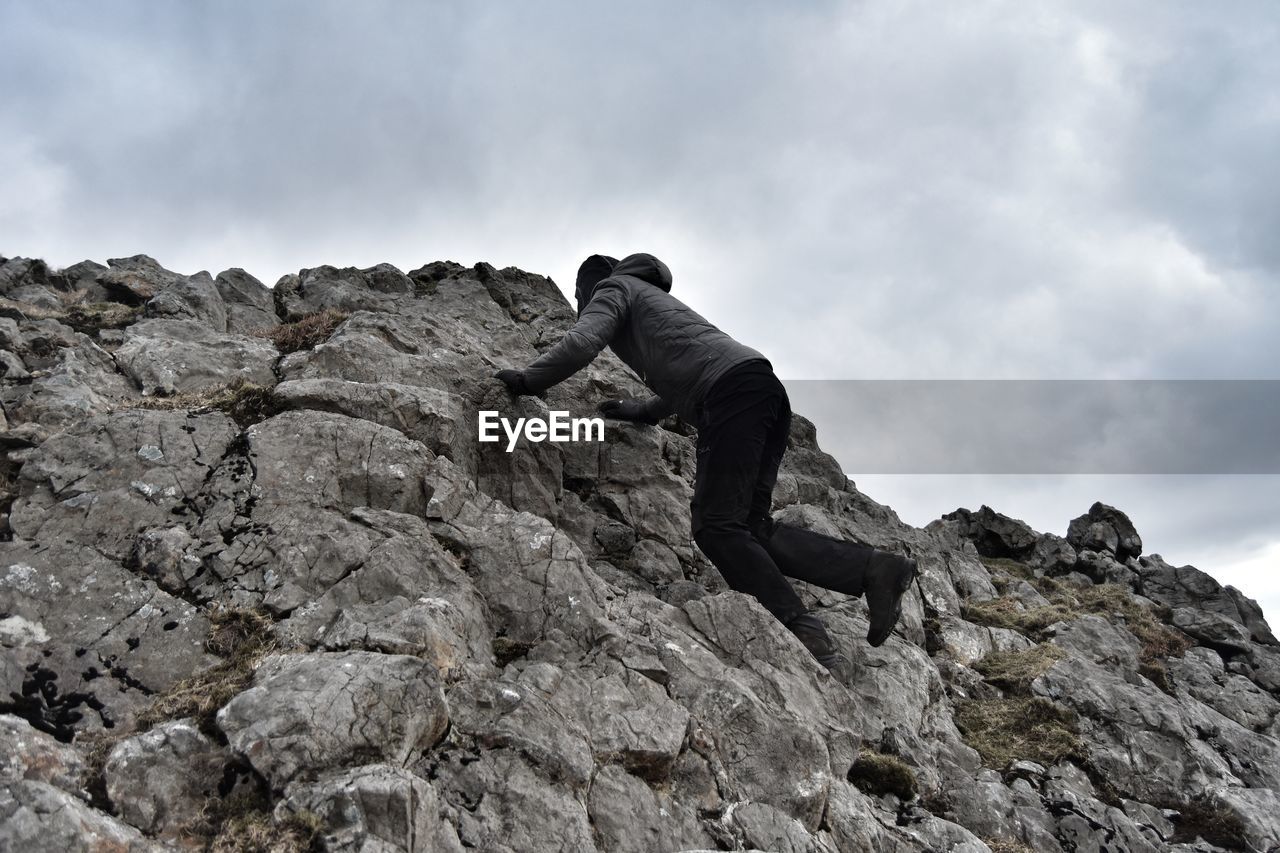 Low angle view of man climbing on rock formation against cloudy sky