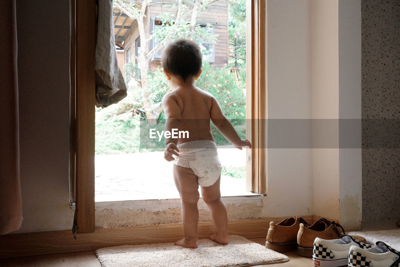 one person, full length, childhood, child, indoors, window, rear view, standing, shorts, day, door, domestic room, entrance, lifestyles, nature, person, domestic life, human leg, home interior, sunlight, men, clothing, doorway, interior design, looking, barefoot, house, room, vacation, leisure activity, trip