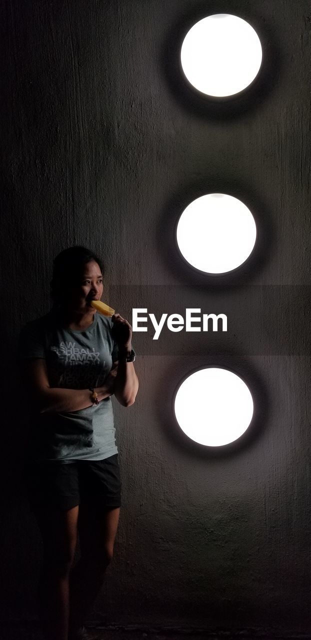 FULL LENGTH OF YOUNG WOMAN STANDING AGAINST ILLUMINATED CEILING