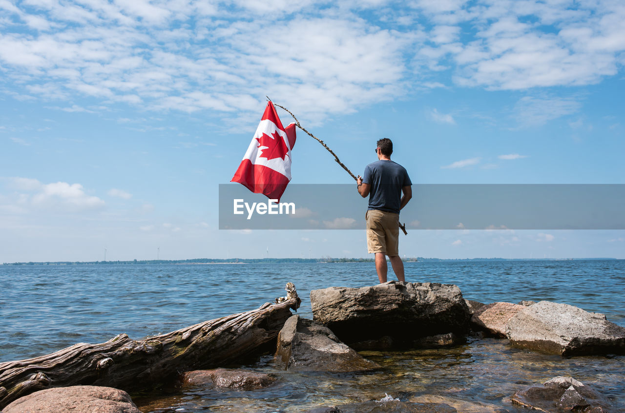 Man holding canada flag on rocky shore of a lake on a summer day.