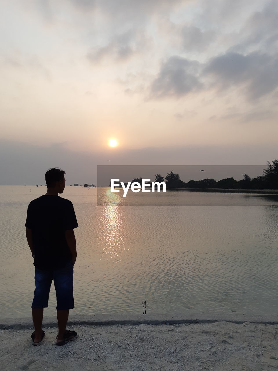 REAR VIEW OF MAN STANDING ON SHORE AGAINST SKY DURING SUNSET