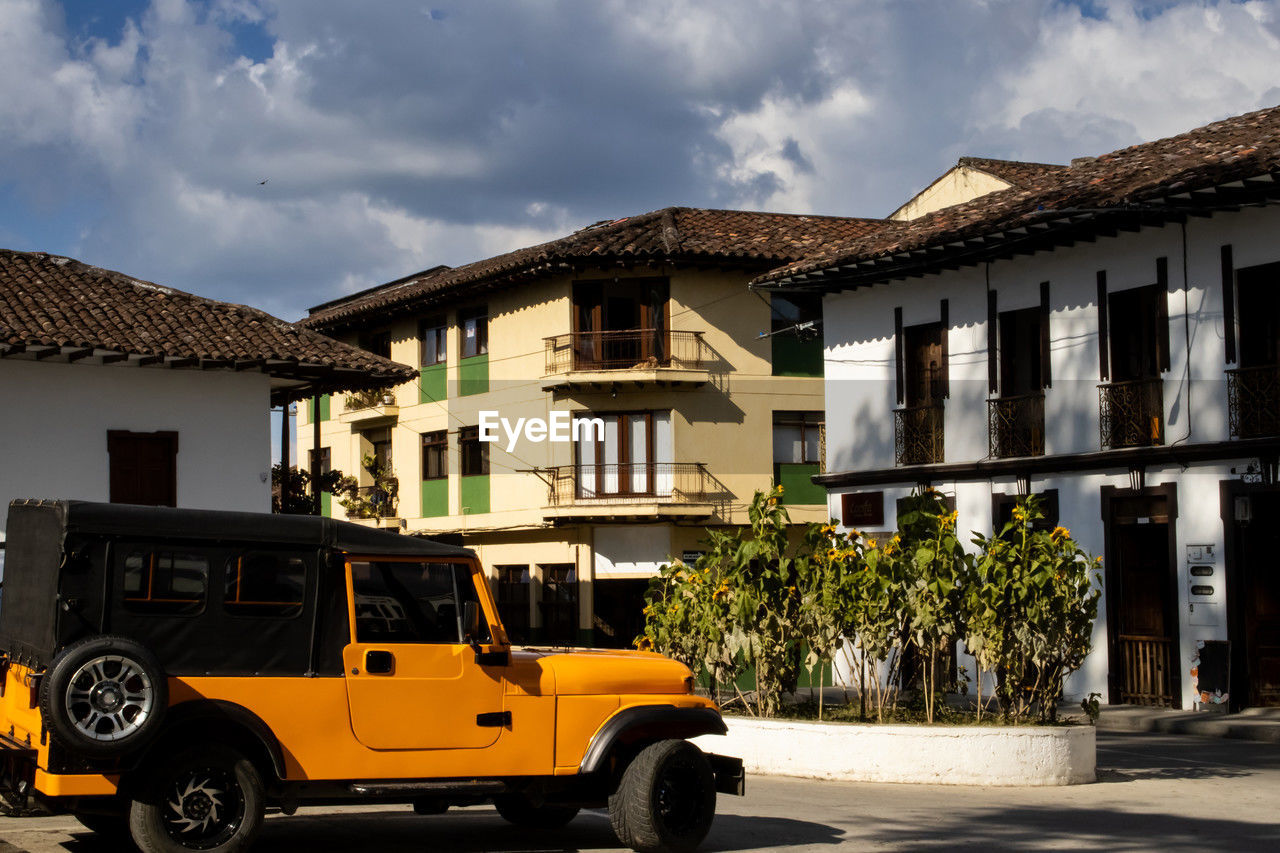 architecture, building exterior, transportation, mode of transportation, car, built structure, building, vehicle, city, motor vehicle, residential district, street, house, land vehicle, cloud, nature, sky, yellow, road, outdoors, travel, wheel, no people, city street