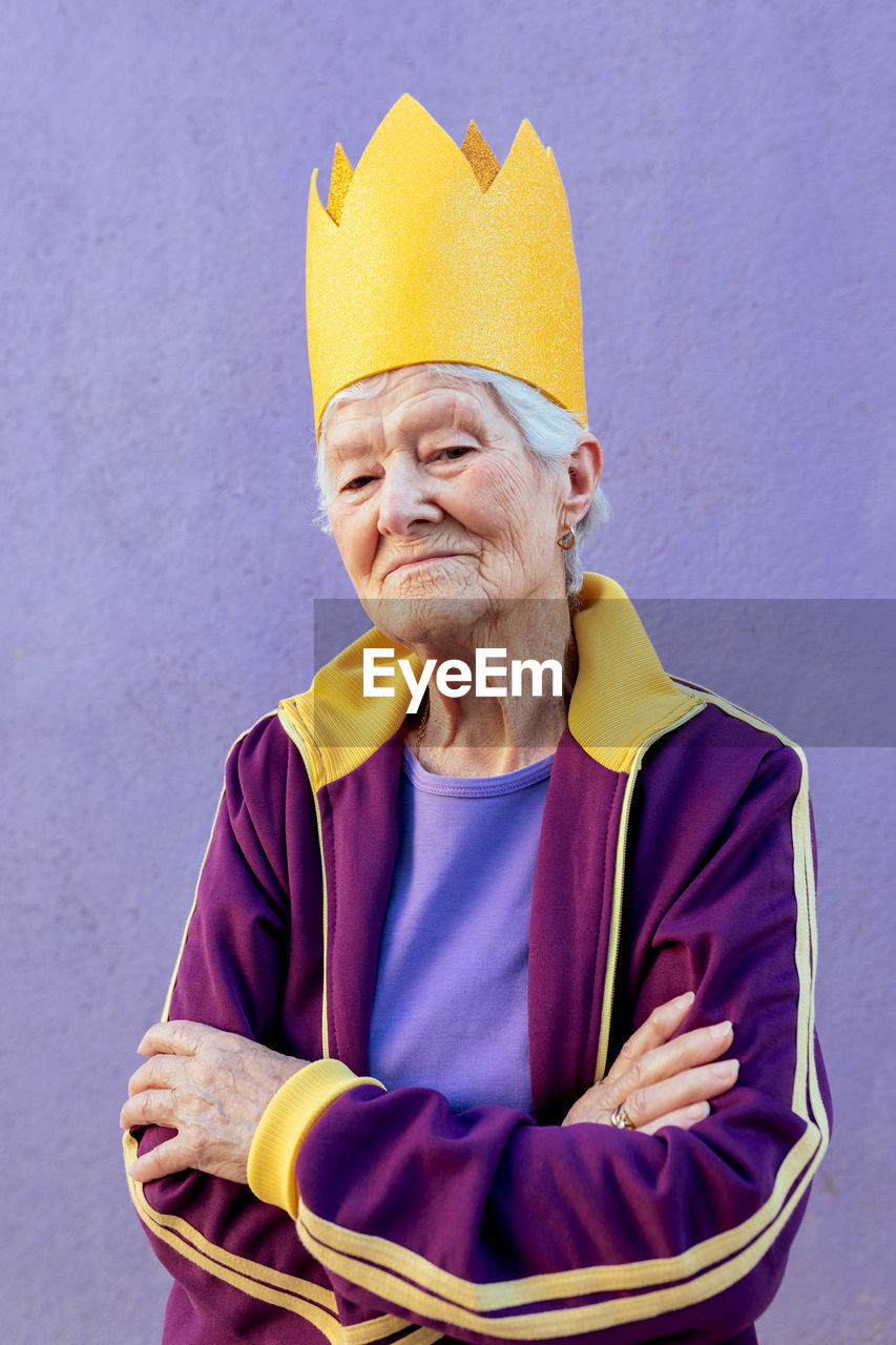 Confident elderly female athlete in sportswear and decorative crown looking at camera with folded arms on purple background