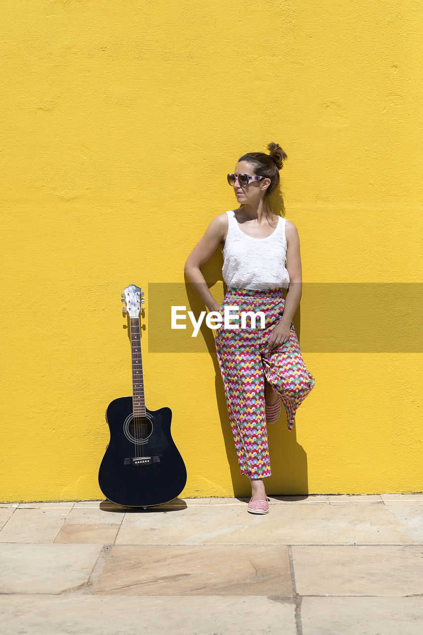 Pretty guitarist woman looking away with guitar leaning on yellow wall