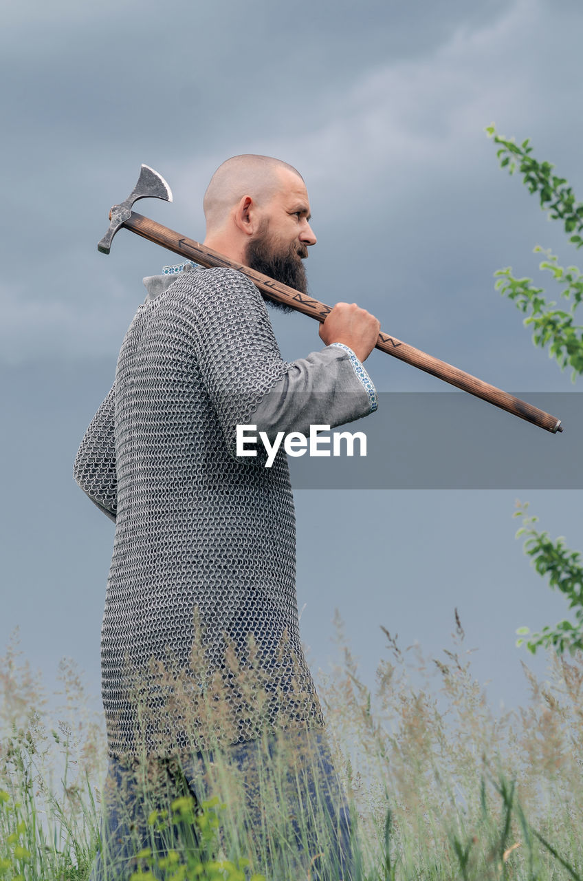 one person, adult, men, plant, nature, standing, sky, holding, grass, leisure activity, day, three quarter length, beard, facial hair, side view, mature adult, weapon, land, looking, rural scene, clothing, casual clothing, outdoors, activity, waist up, landscape, field