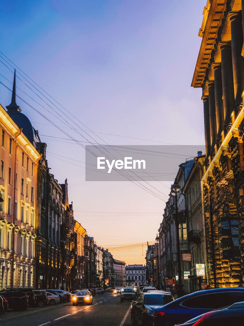 CARS ON STREET AMIDST BUILDINGS IN CITY AT SUNSET