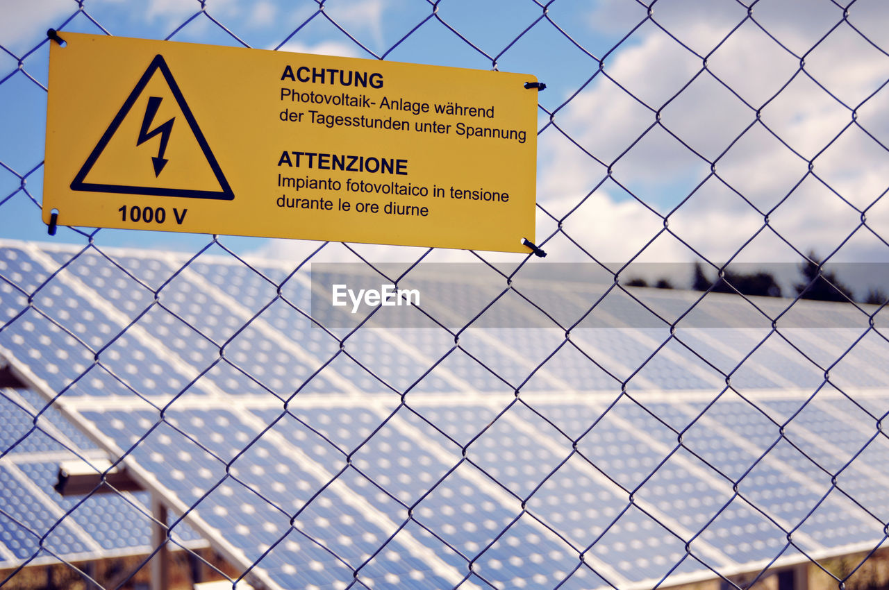 Close-up of high voltage sign on fence against solar panels